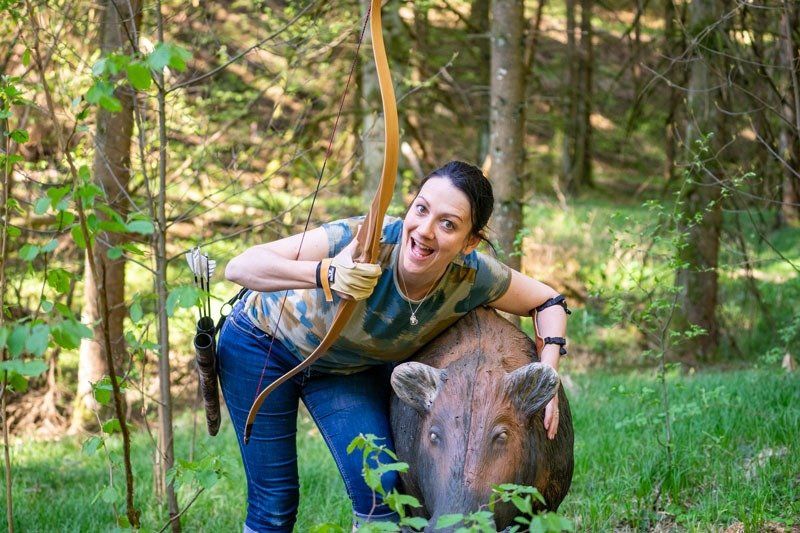 Eine Frau in Tarnkleidung spannt einen Bogen und lächelt neben einer Wildschweinzielscheibe im Wald.