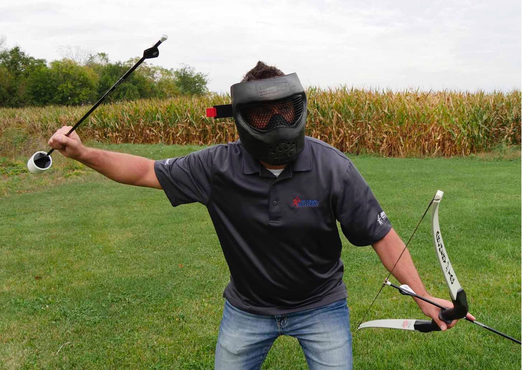 Ein Mann mit Paintballmaske zielt im Freien vor einem Maisfeld mit Pfeil und Bogen.