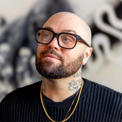 Bald man with glasses and beard wearing a dark sweater and gold chains against a blurred black-and-white background