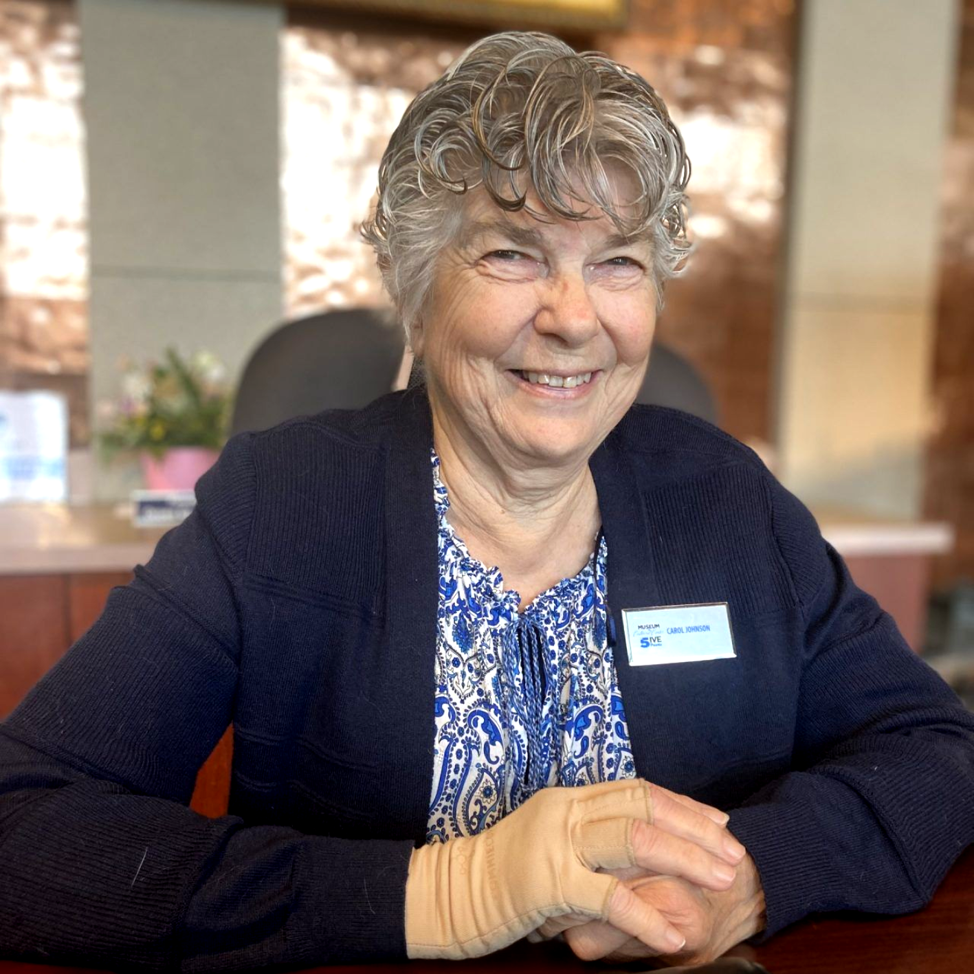 Smiling person seated indoors at a table, wearing a navy cardigan and name tag, with bandaged hands.