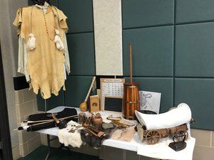 Display of vintage clothing, books, and crafts on a table with a hanging fringed garment against a tiled wall.