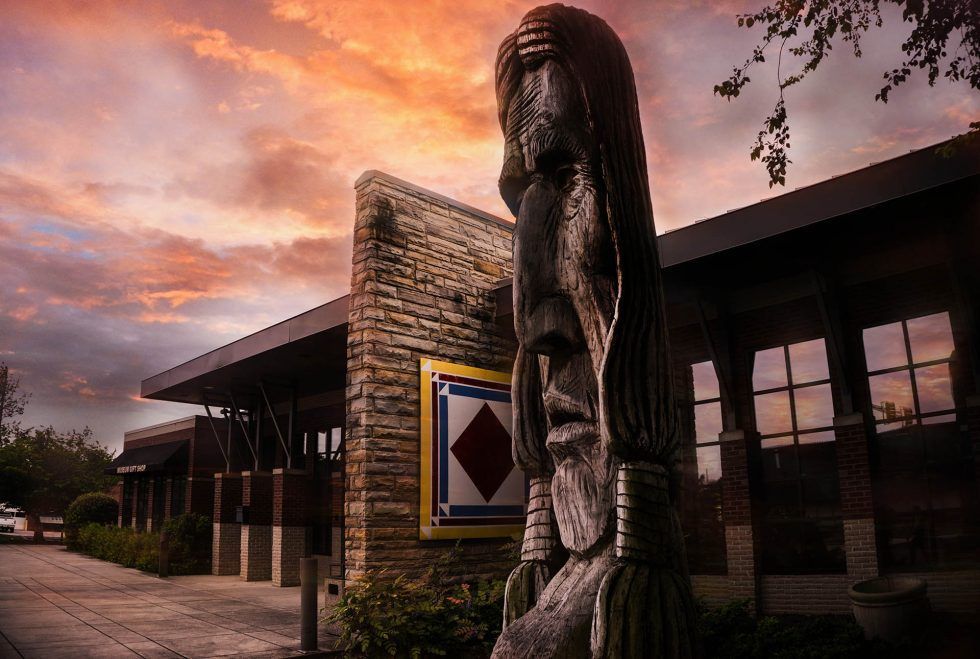Wooden totem beside a brick building at sunset, with pink clouds and windows reflecting the evening light
