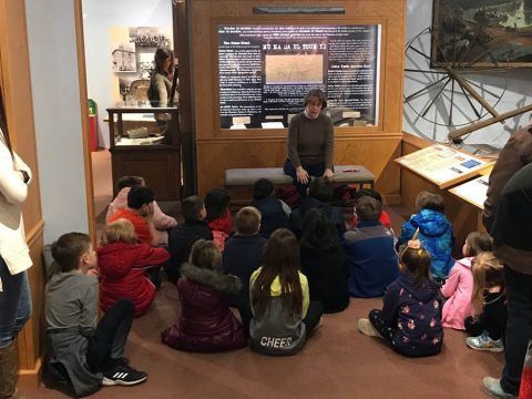 Children seated on the floor listen to a speaker in a classroom with a chalkboard display.