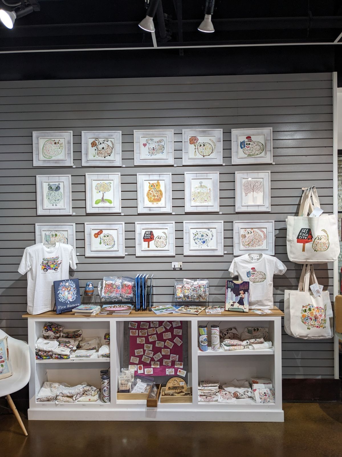 Merchandise display with framed art, shirts, tote bags, and gifts against a gray slat wall