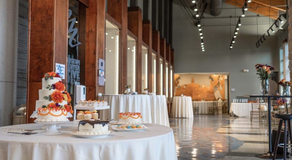 Elegant banquet hall with round white-clothed tables, tall windows, and a decorated dessert display.
