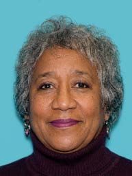 Studio headshot of a smiling woman in a black blazer and pearl necklace on a gray background
