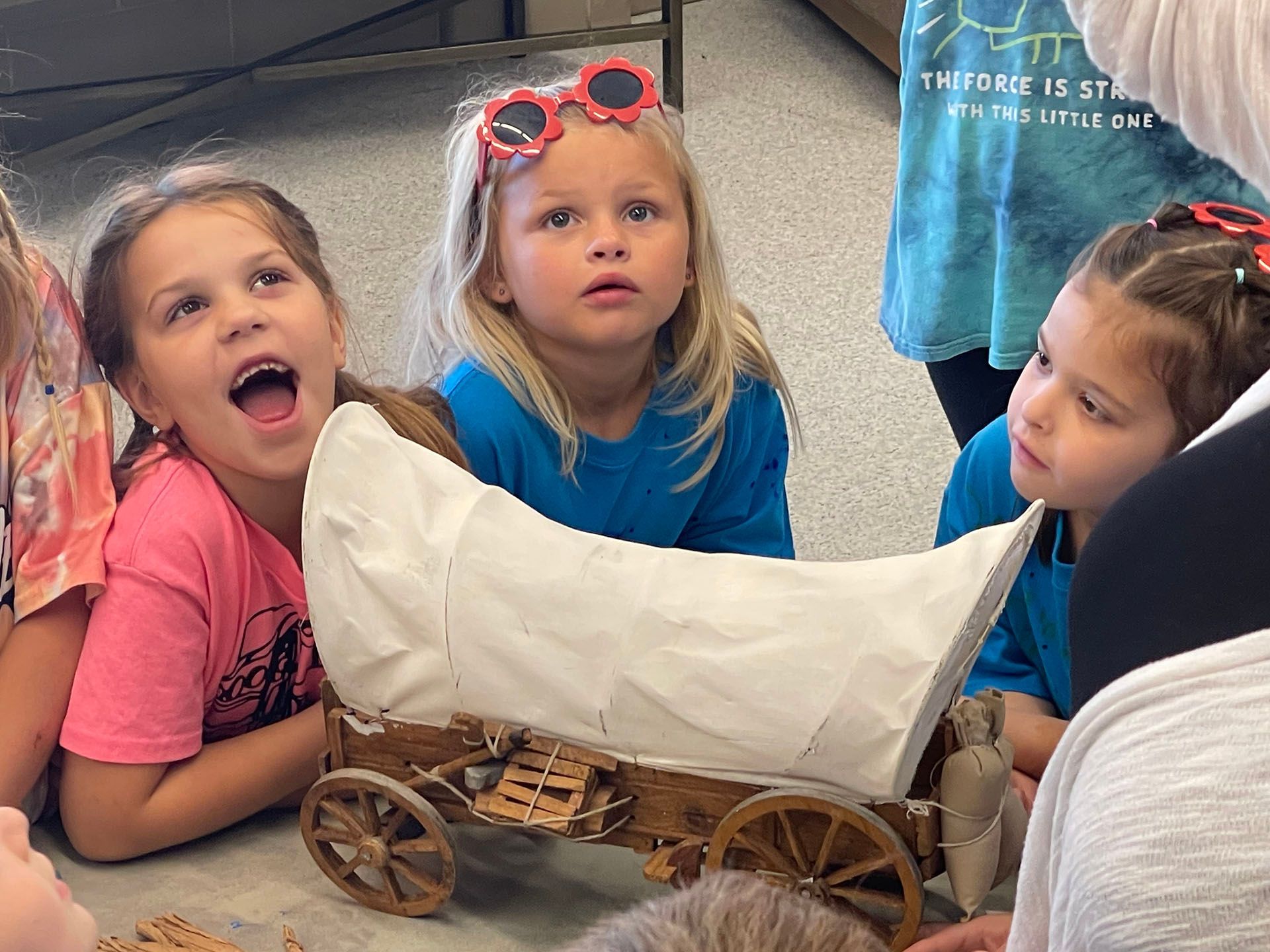 Three children in colorful shirts gather around a small wagon indoors, looking at its white cloth cover.