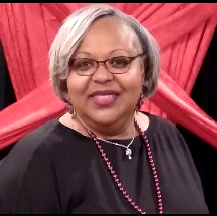Studio headshot of a smiling woman in a black blazer and pearl necklace on a gray background