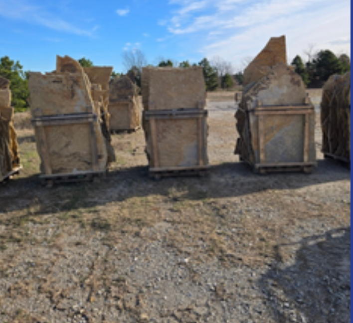 Several large, flat, beige stone slabs stand upright in wooden shipping crates on a gravel lot under a blue sky.