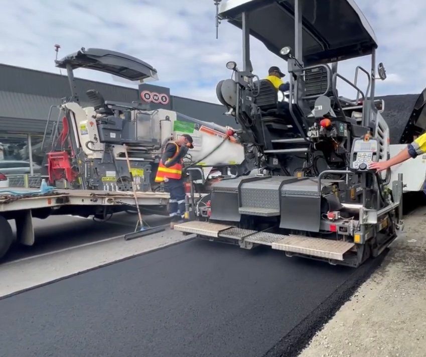 Road paving machine laying fresh black asphalt while workers operate nearby.