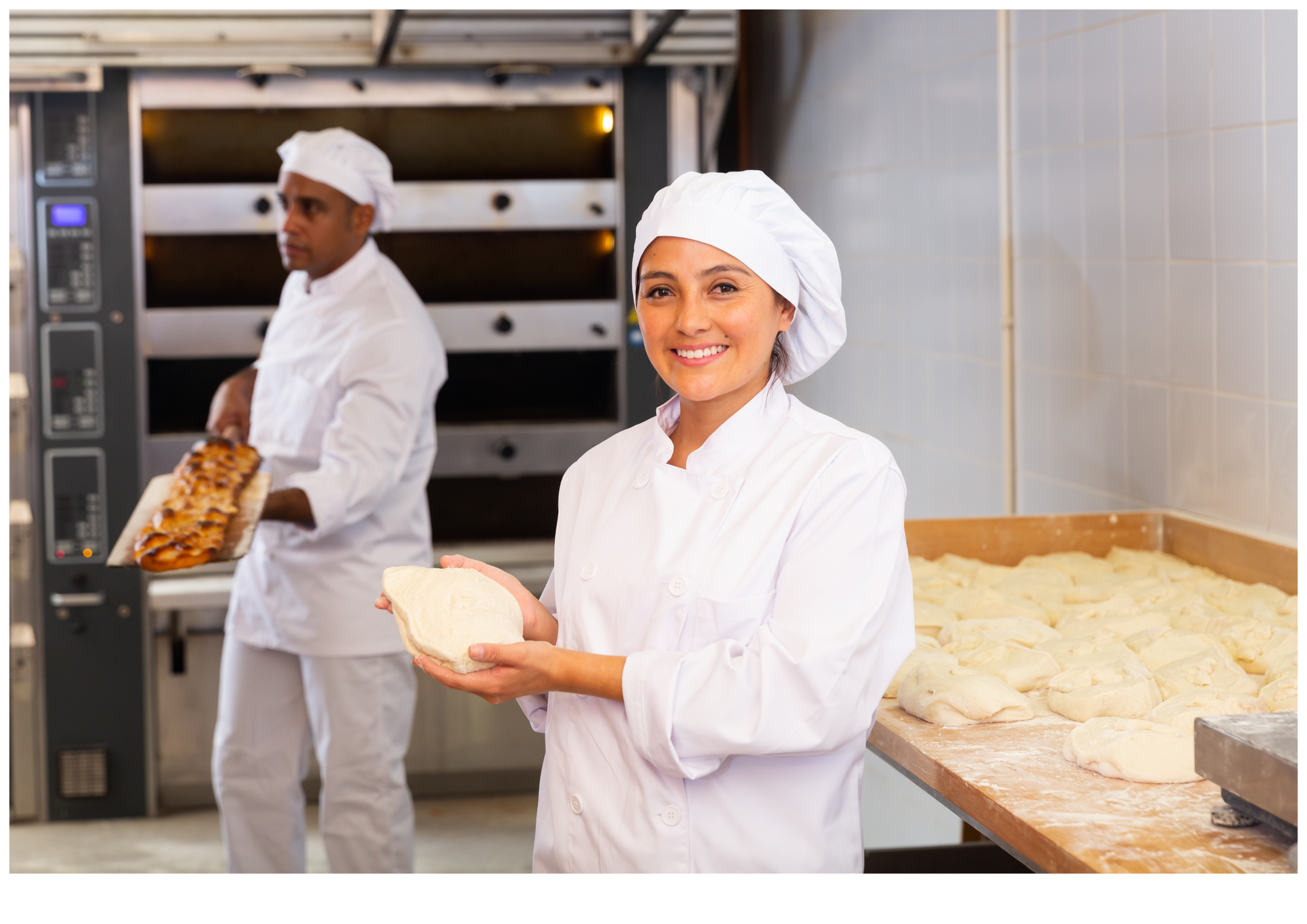Bakers in white uniforms in a bakery, one smiling while holding dough and trays of bread in the background
