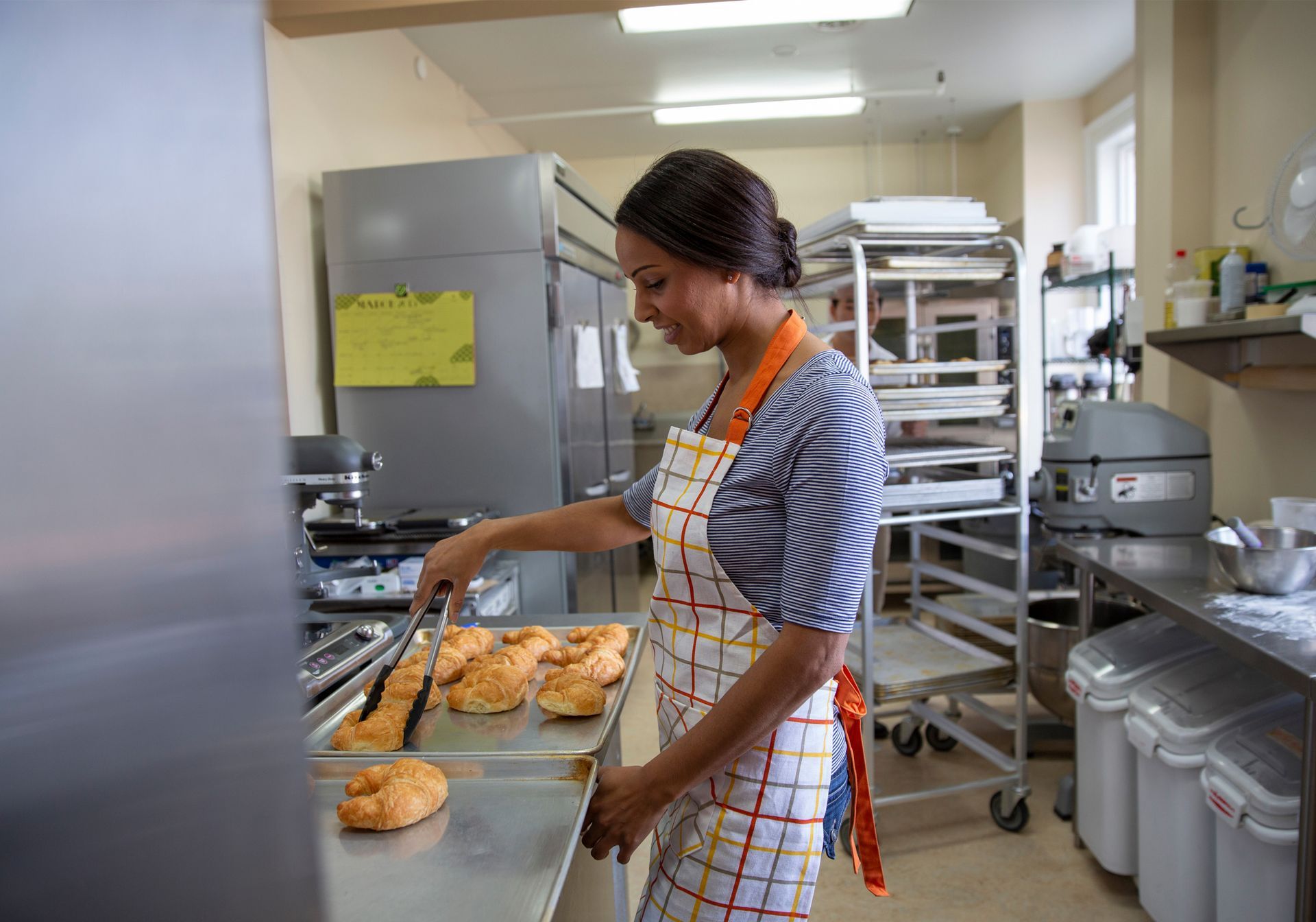 Worker in an industrial kitchen placing pastries on trays beside a stainless-steel counter