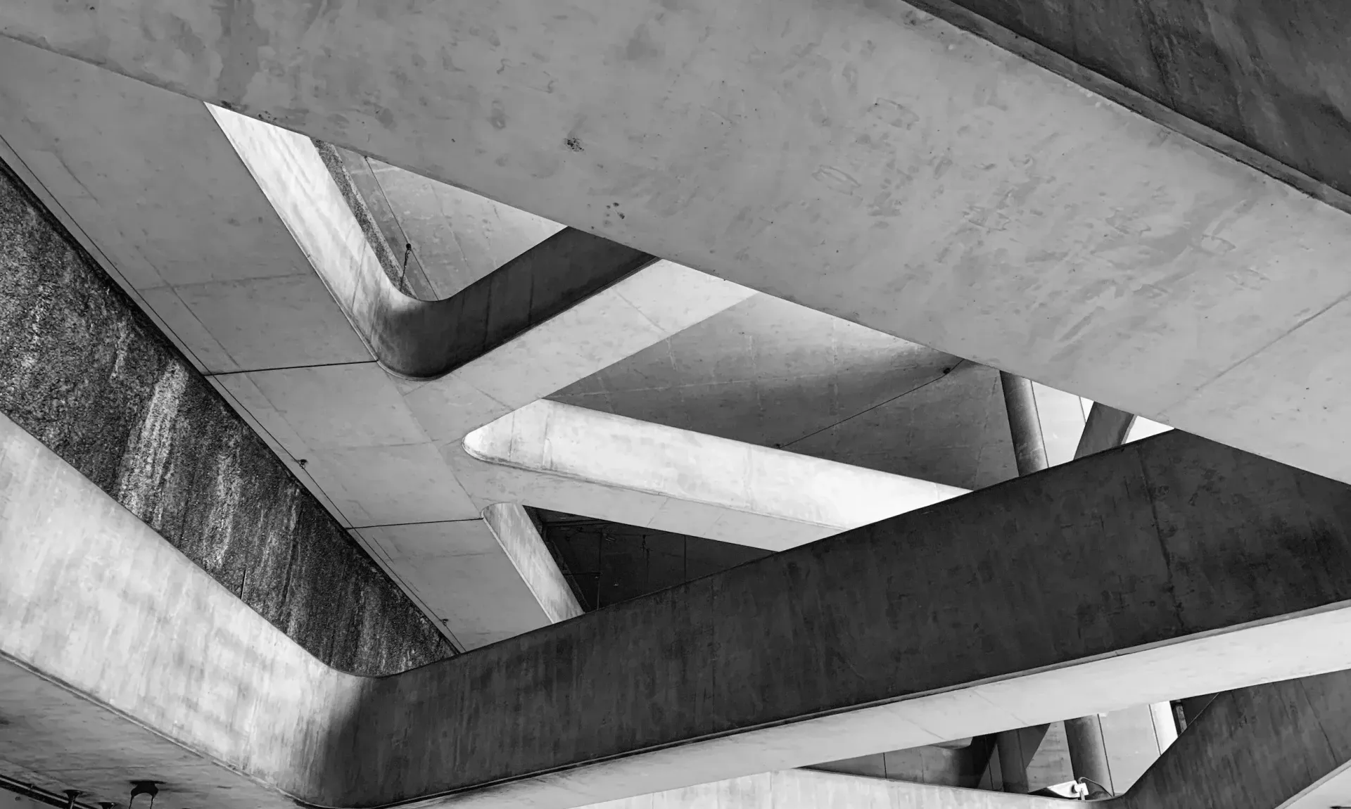 Monochrome view of intersecting metal beams and concrete supports under a bridge or structure