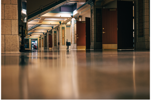 Empty indoor corridor with glossy floor, pillars, and overhead stairs in soft warm light