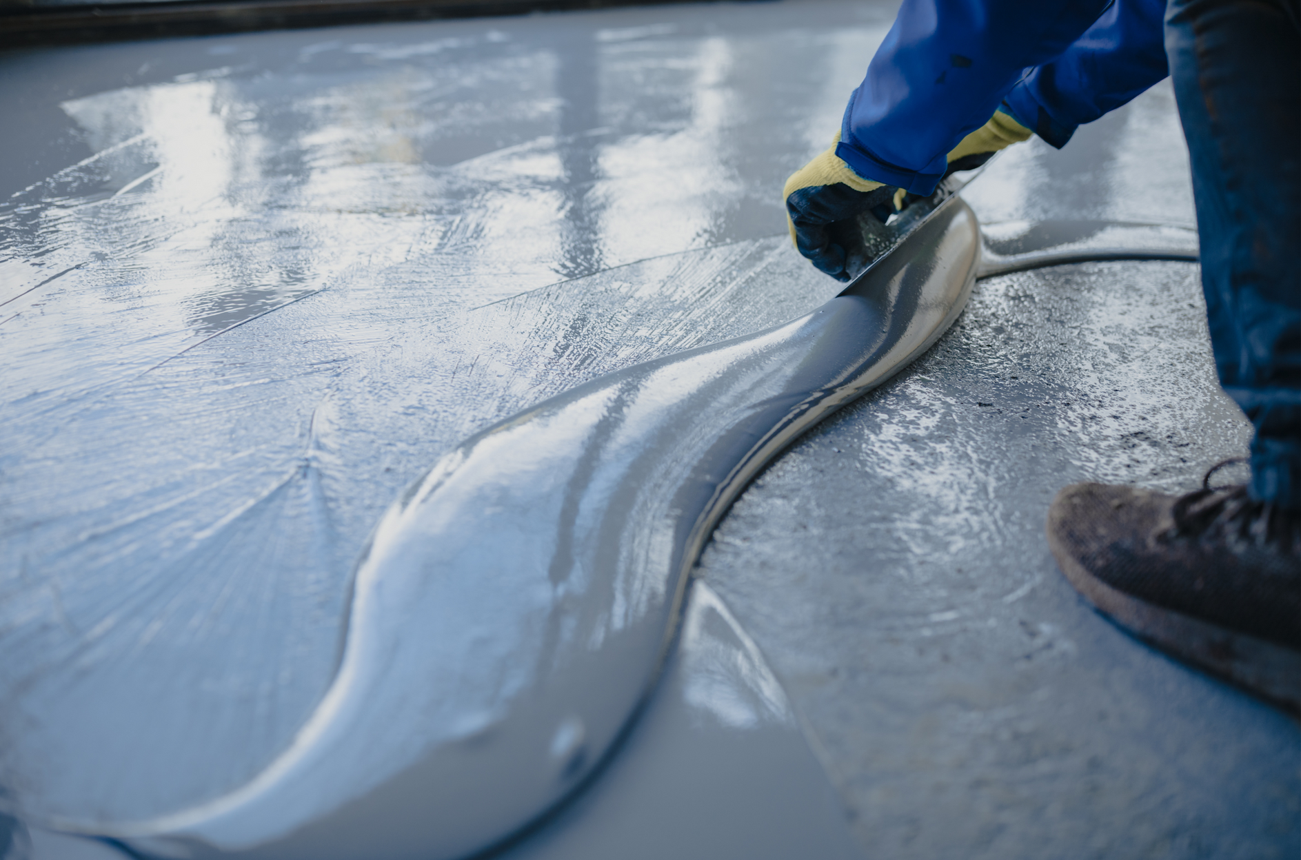 Worker smoothing wet concrete floor with a trowel, wearing blue gloves and boots
