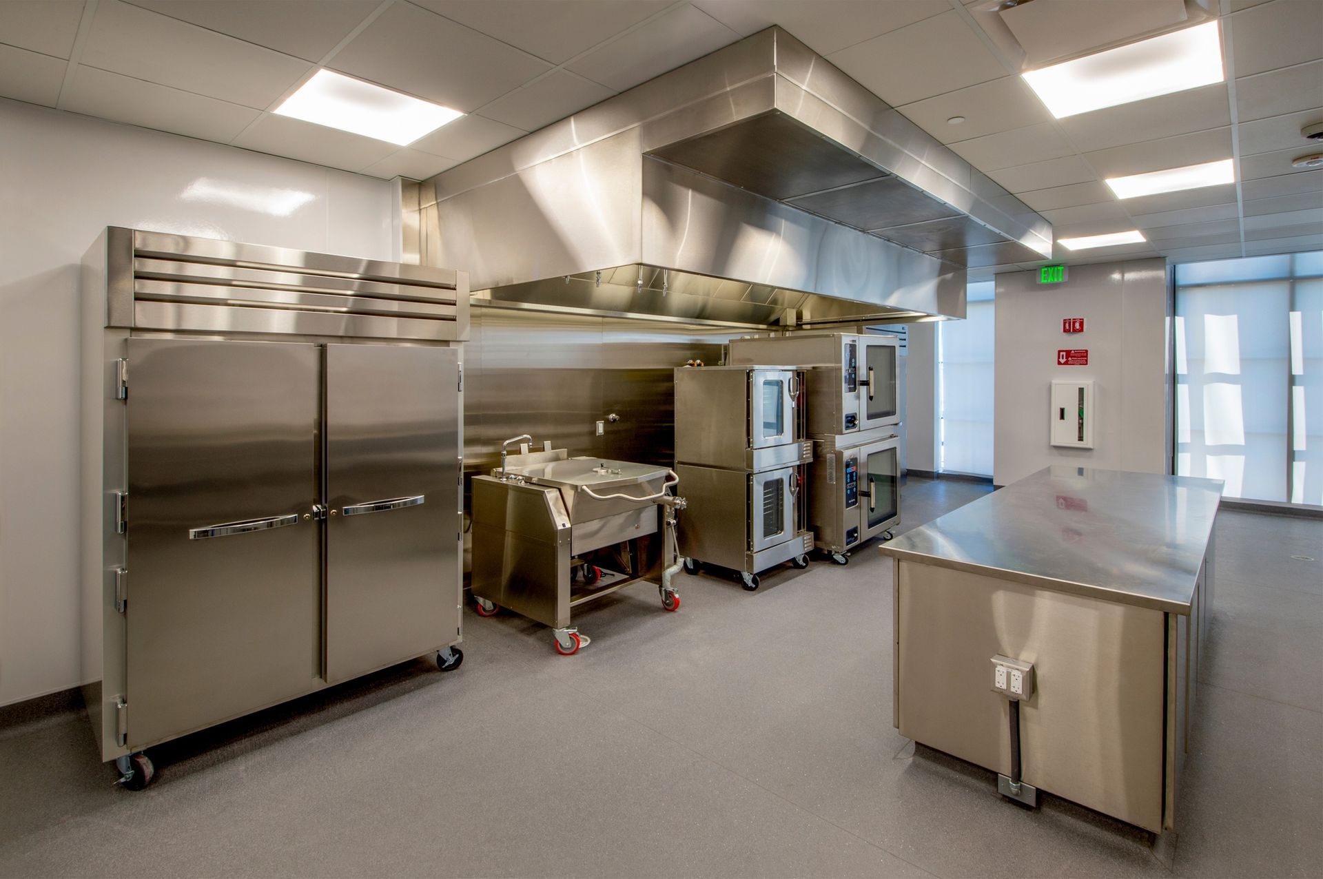 Stainless steel commercial kitchen with ovens, prep tables, and ventilation hood under bright ceiling lights