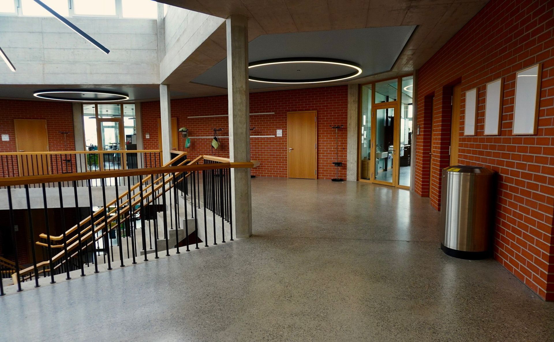 Indoor school hallway with brick walls, railing, bulletin boards, and a trash can.
