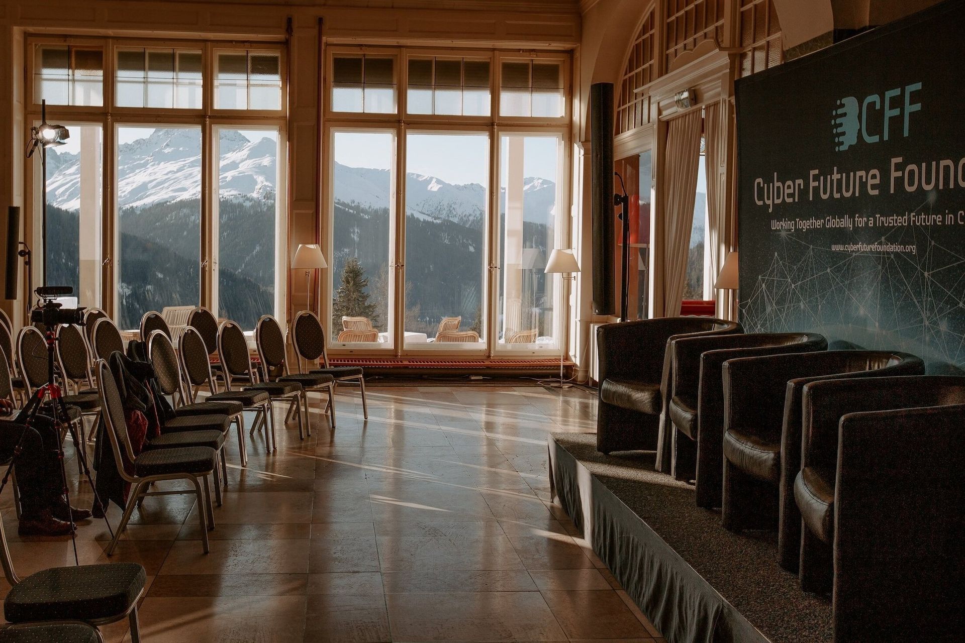 Empty conference hall with rows of chairs, a stage, and large windows overlooking mountains.