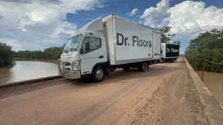 Two white Dr. Floors trucks driving across a dirt bridge over a river under a cloudy sky