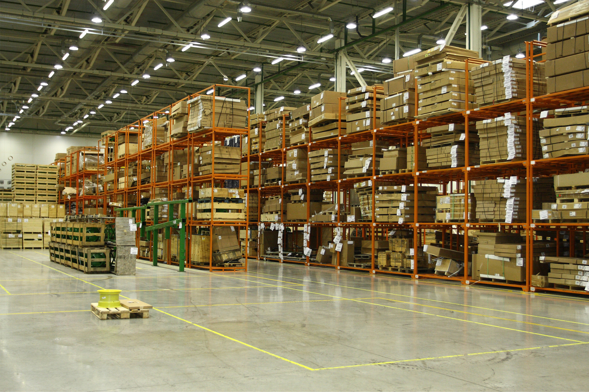 Large warehouse aisle with tall orange racks and stacked pallets under bright overhead lights