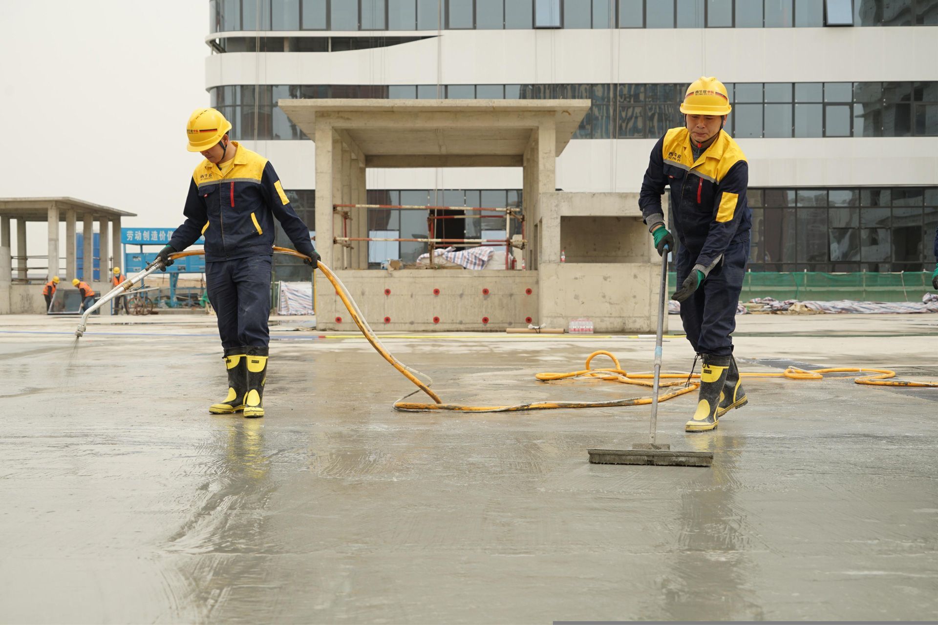 Two workers in yellow hard hats hose down a wet concrete surface outside a building