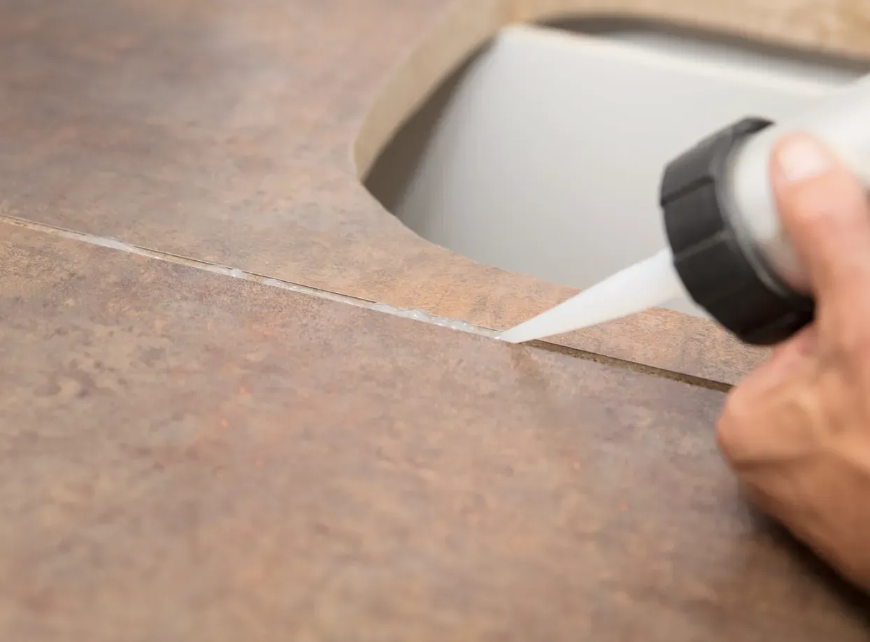 A hand applies white sealant to a crack in a brown countertop near a sink.