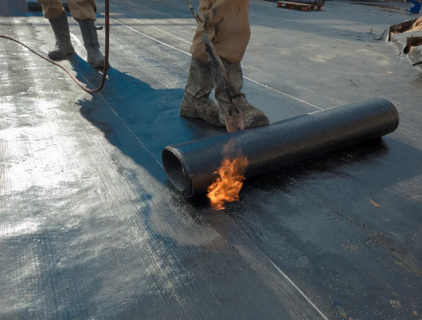 Worker torch-sealing a black roofing membrane on a flat roof