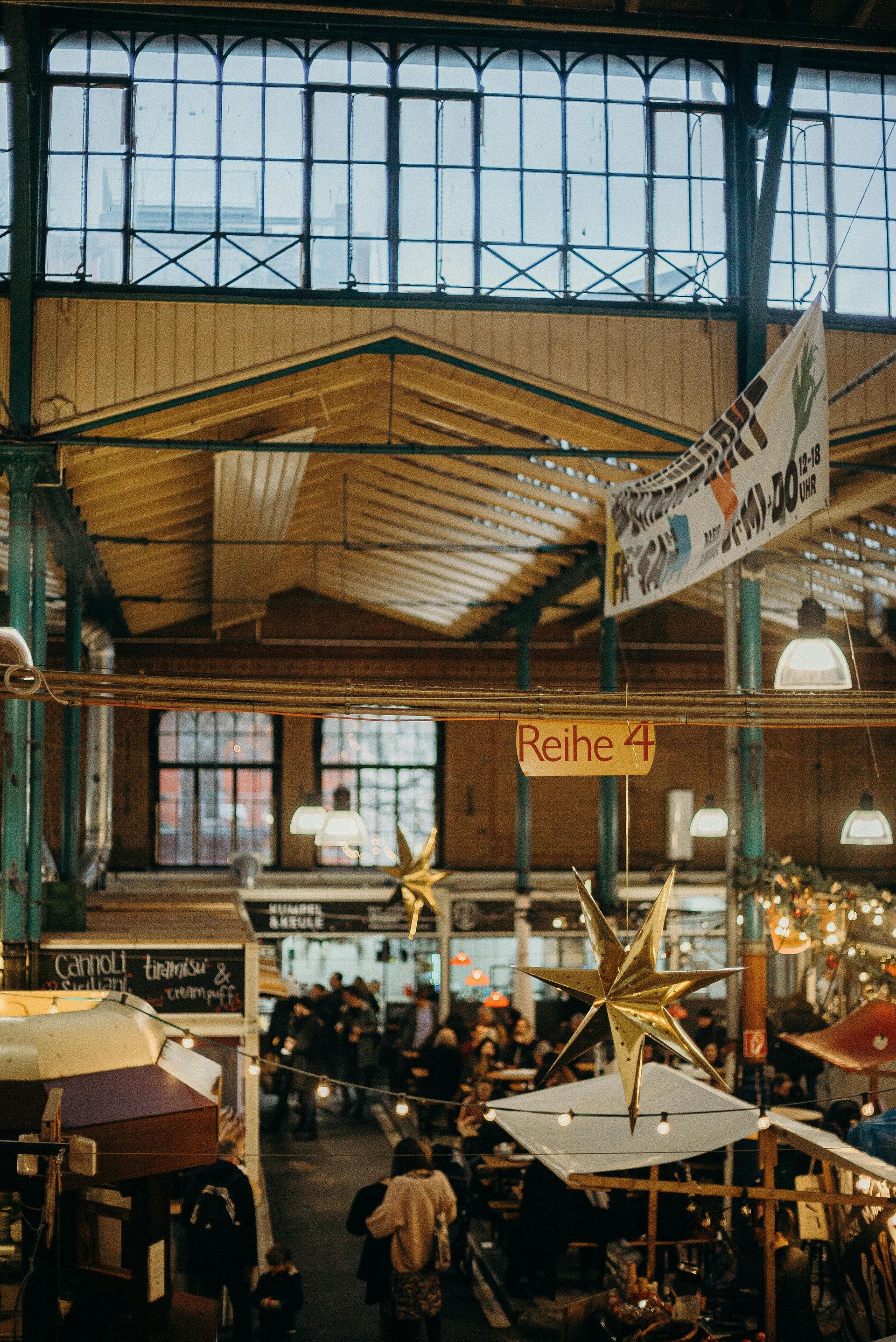 Bustling indoor market with tall windows, hanging signs, and crowded stalls under a wooden roof