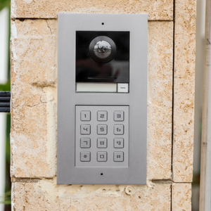 Silver intercom with keypad, camera, and speaker mounted on a stone wall.