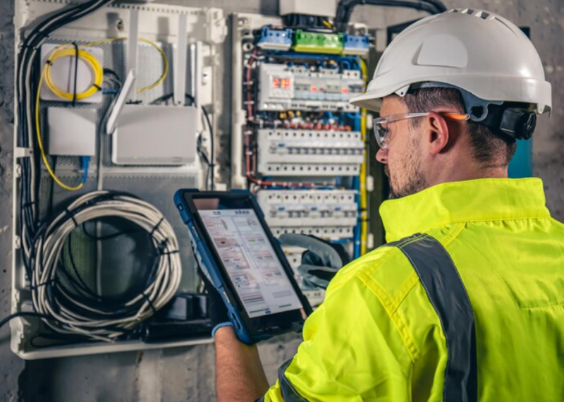 Electrician in hard hat using a tablet to inspect wiring inside an open electrical panel