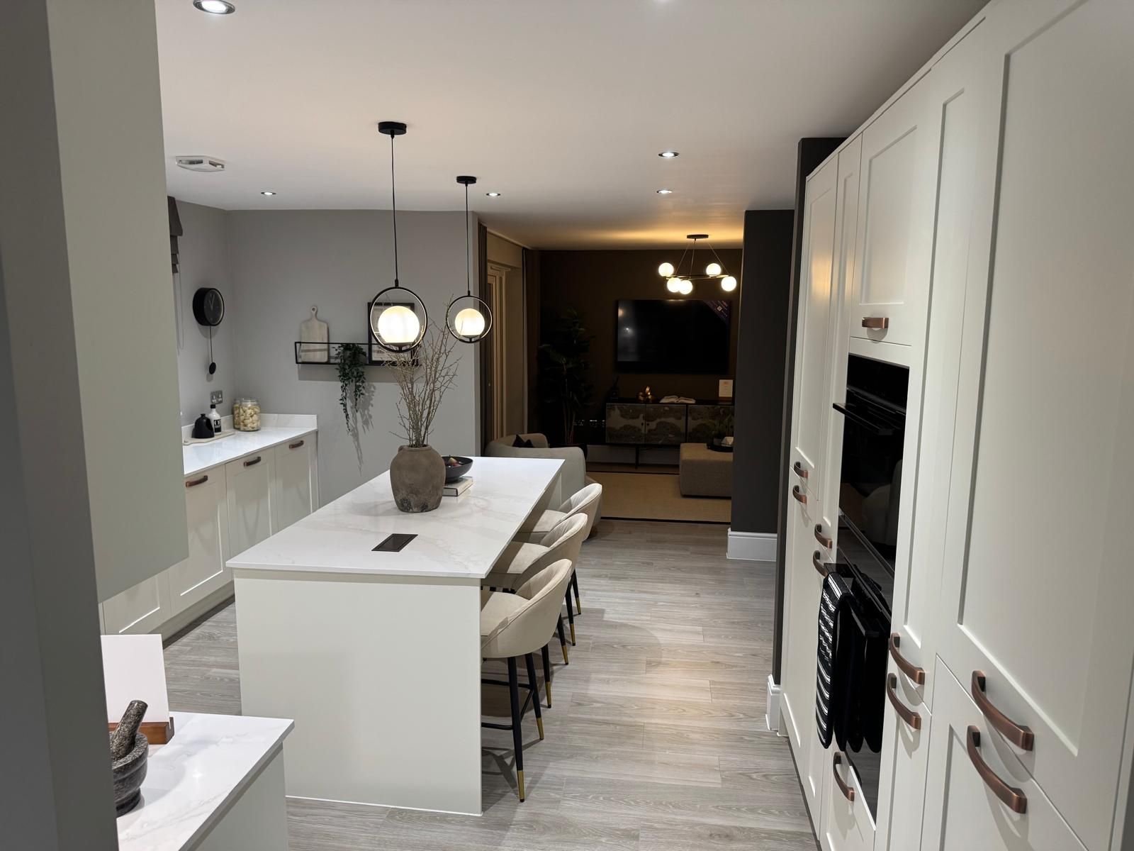 Modern kitchen with island, pendant lights, and built-in ovens, leading to a living room.