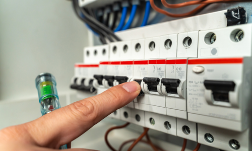 Person's finger pushing a circuit breaker switch in a white electrical panel. A light tester glows green.