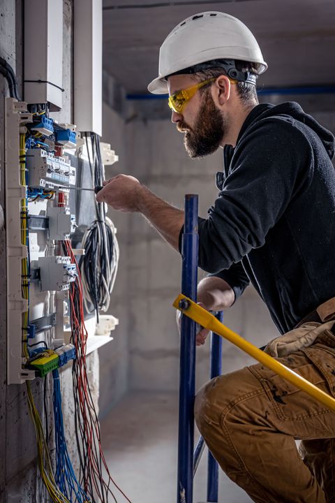 Electrician in hardhat and safety glasses works on electrical panel.
