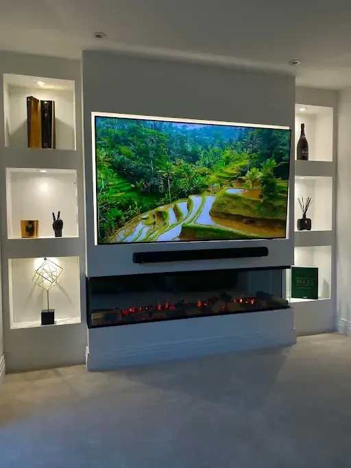 Built-in media wall unit in Fareham  with large TV, fireplace, and side shelving. Green landscape on screen, white surround.