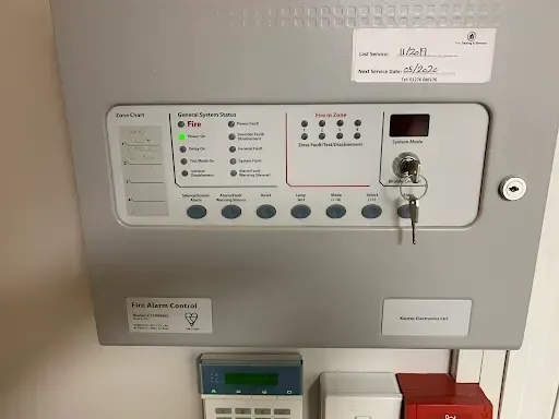 Fire alarm control panel on a gray wall with various indicator lights and a keypad.
