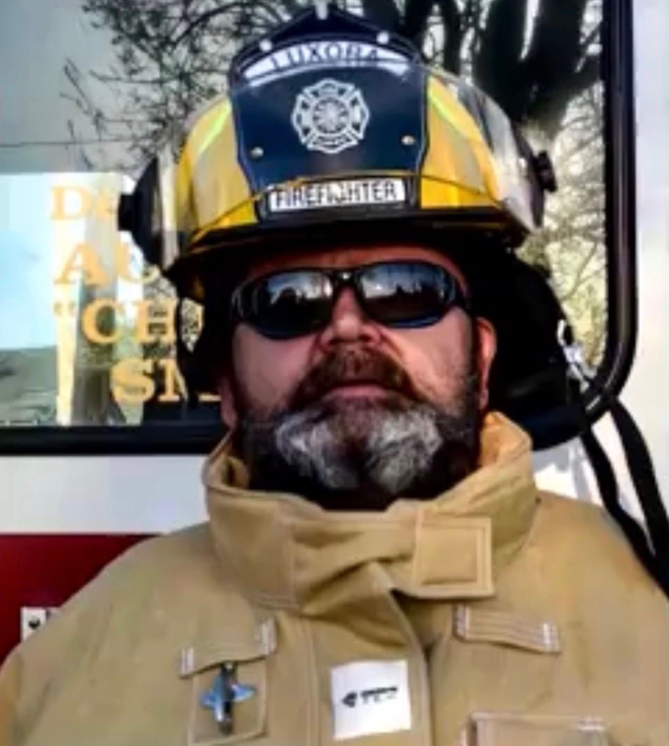 Firefighter in tan turnout gear and helmet, wearing sunglasses and a beard, standing beside a fire truck.