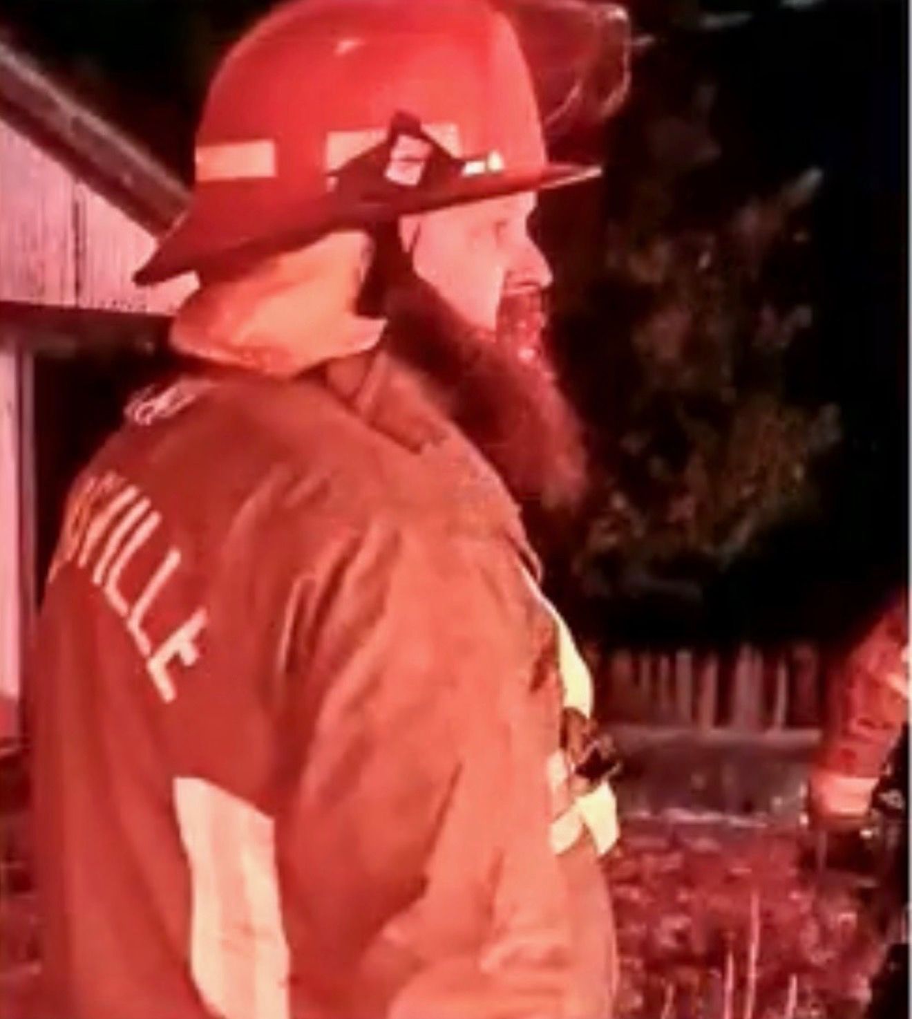 Firefighter in red helmet and brown jacket standing outdoors at night near a house, lit by red emergency light