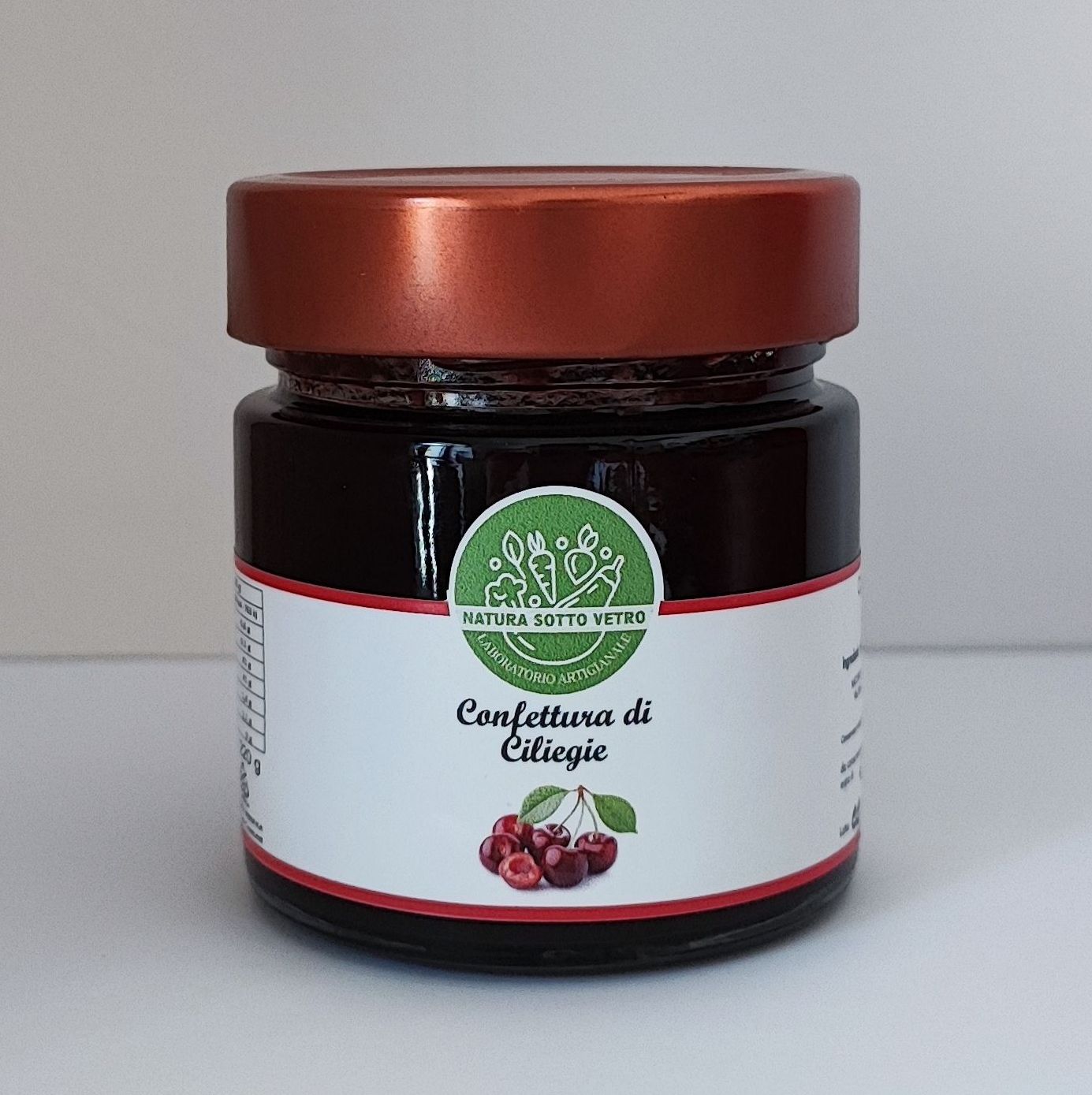 Vasetto di marmellata di ciliegie con coperchio color rame ed etichetta bianca su sfondo neutro.