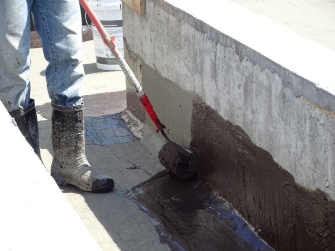 Worker applying black waterproof coating to a concrete wall with a roller on a rooftop.