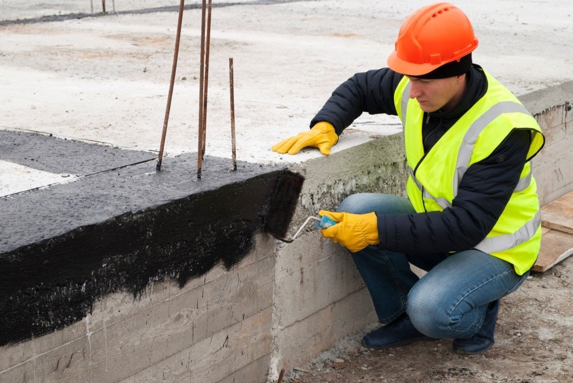 Construction worker in orange hard hat applying sealant to a concrete curb at a roadside worksite