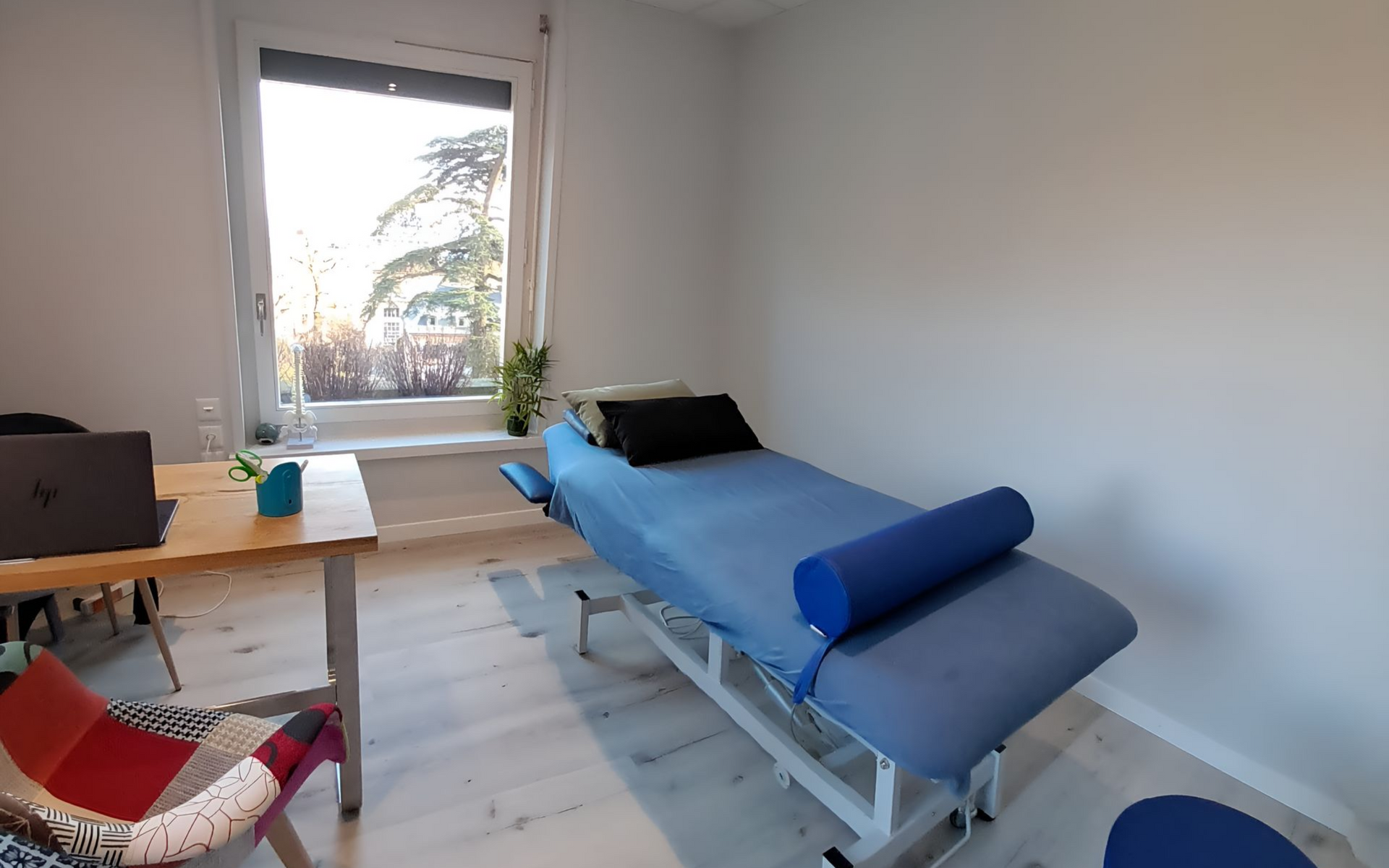 Salle de thérapie avec une table de massage bleue, un petit bureau avec un ordinateur portable et une fenêtre.