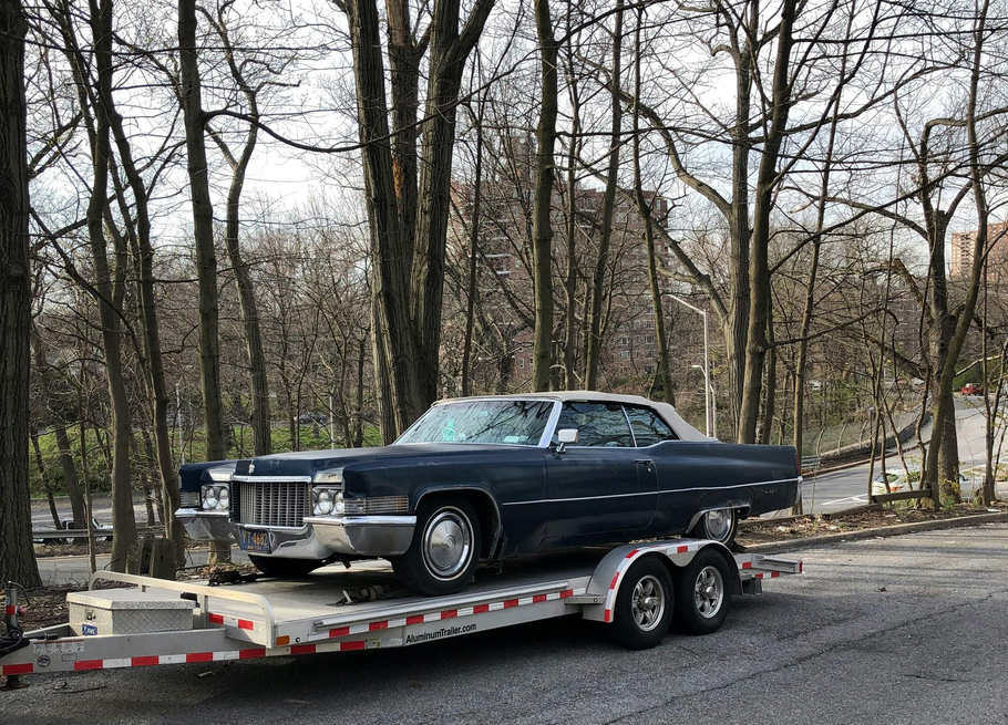 A dark blue vintage Cadillac on a flatbed trailer parked on a roadside with bare trees in the background.
