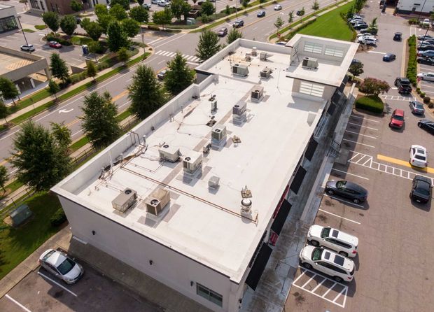 Overhead view of a building with a flat white roof, AC units, and parking lot with cars.