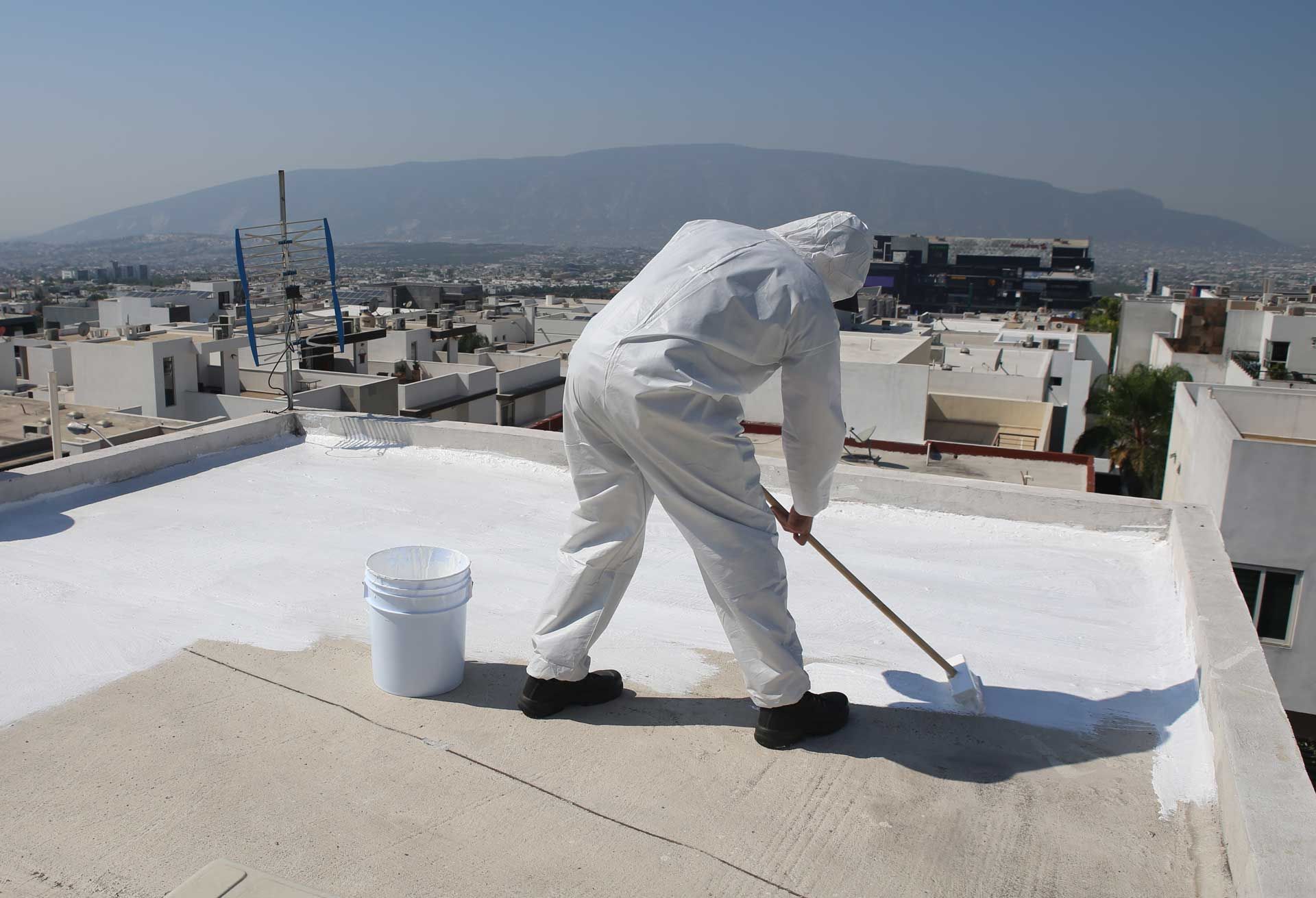 Person in protective suit applying white coating to a flat rooftop with a roller. City and mountain background. Person in protective suit applying white coating to a flat rooftop with a roller. City and mountain background.