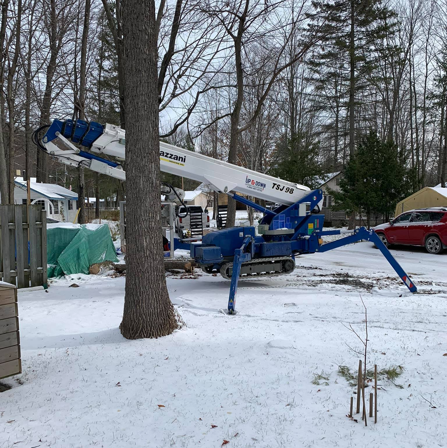 A blue and white aerial lift in a snowy yard, next to a tree, with houses in the background.