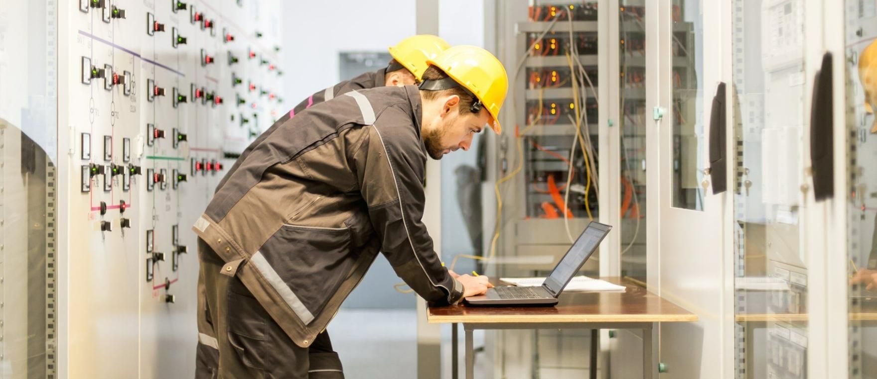 Two workers in yellow hard hats and work clothes using a laptop in a server room.