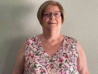 Woman wearing glasses and a floral sleeveless top against a plain gray wall.