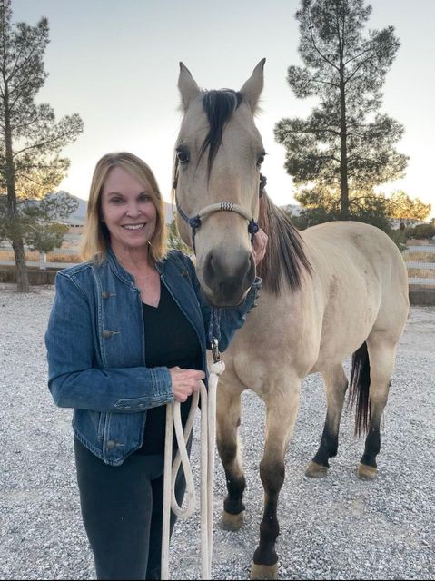 Woman in a denim jacket standing beside a tan horse on a gravel path at sunset