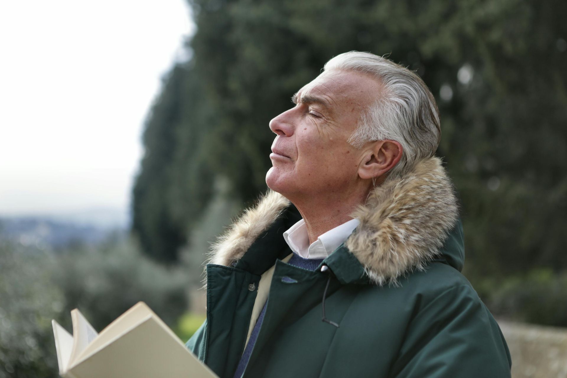 Man in a green coat with fur collar looks upward outdoors while holding an open book