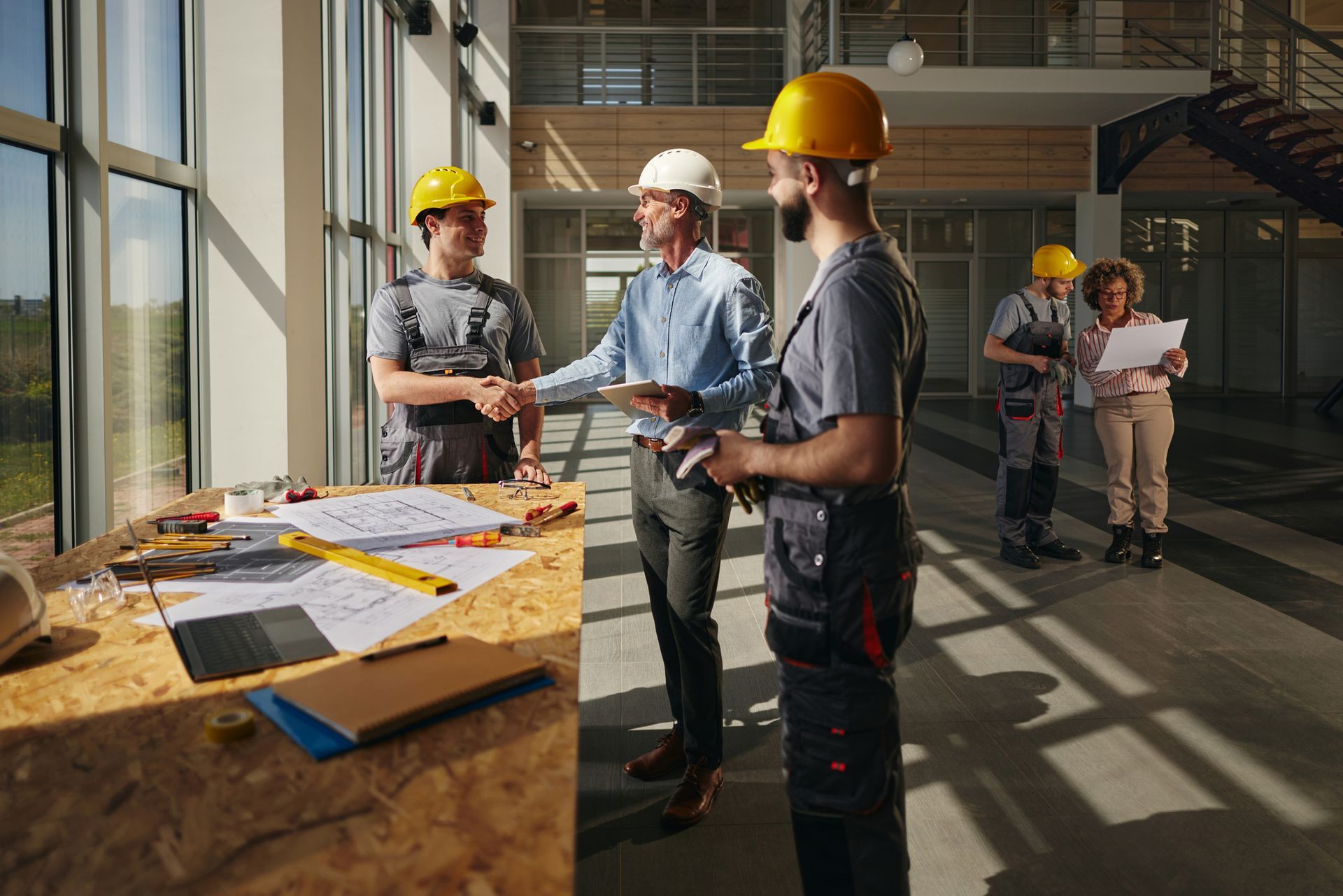 Construction workers and supervisor shaking hands at a worksite. Plans on the table, other workers in the background.